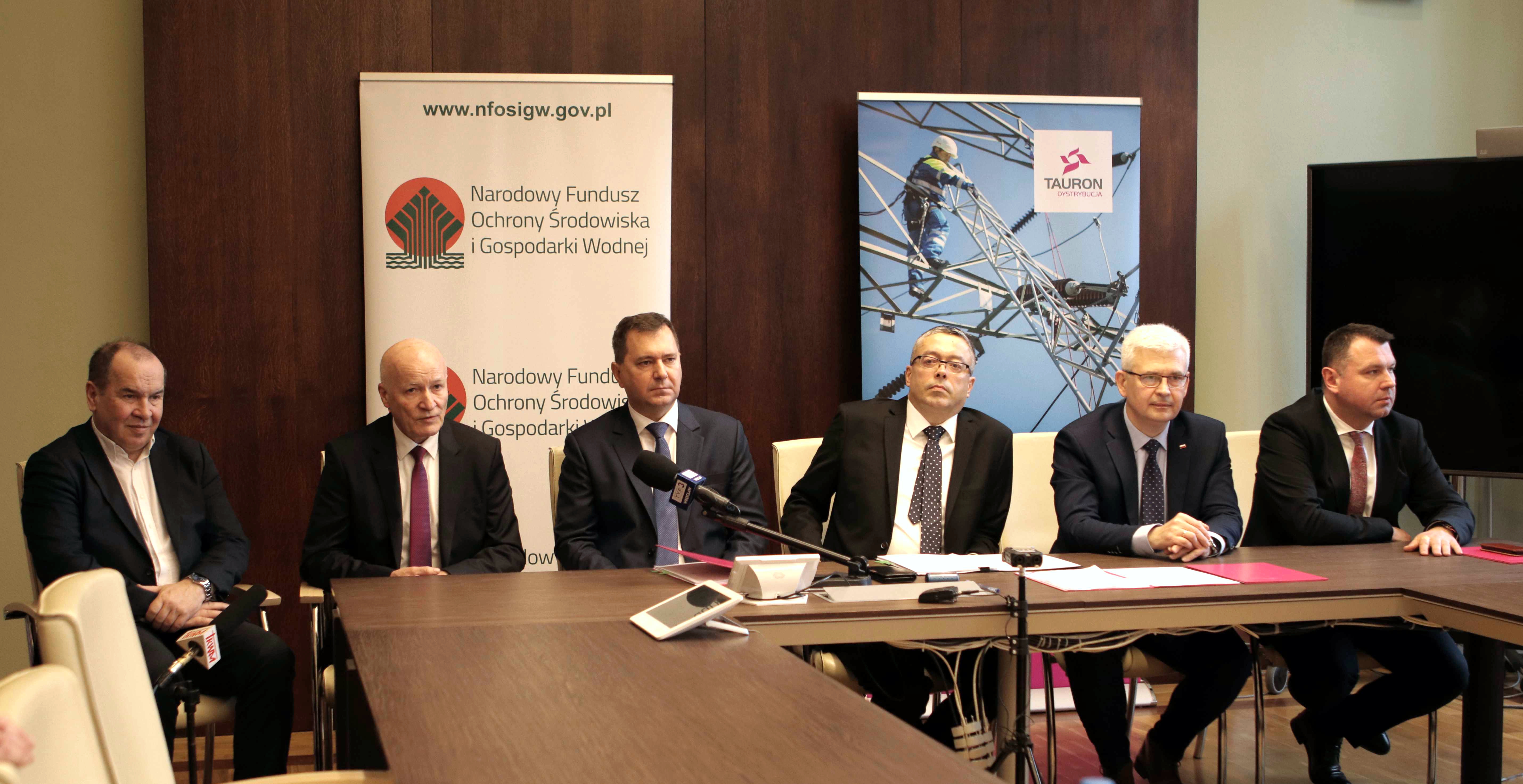 sześciu elegancko ubranych panów siedzi przy stoliku na sali konferencyjnej, za stolikiem są banery Narodowego Funduszu Ochrony Środowiska i Gospodarki Wodnej i firmy TAURON Dystrybucja