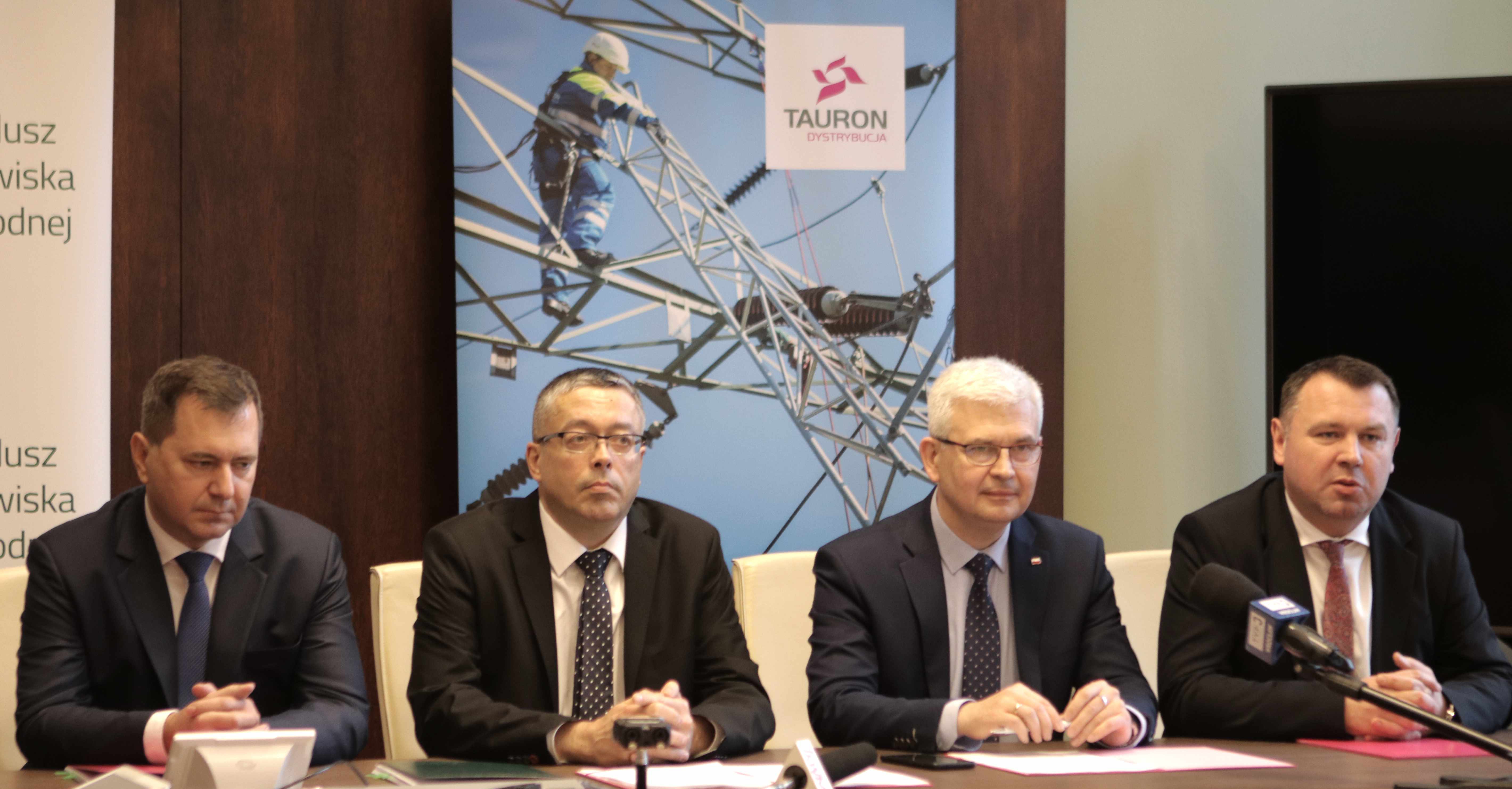 czterech elegancko ubranych panów siedzi przy stoliku na sali konferencyjnej, za stolikiem jest baner firmy TAURON Dystrybucja
