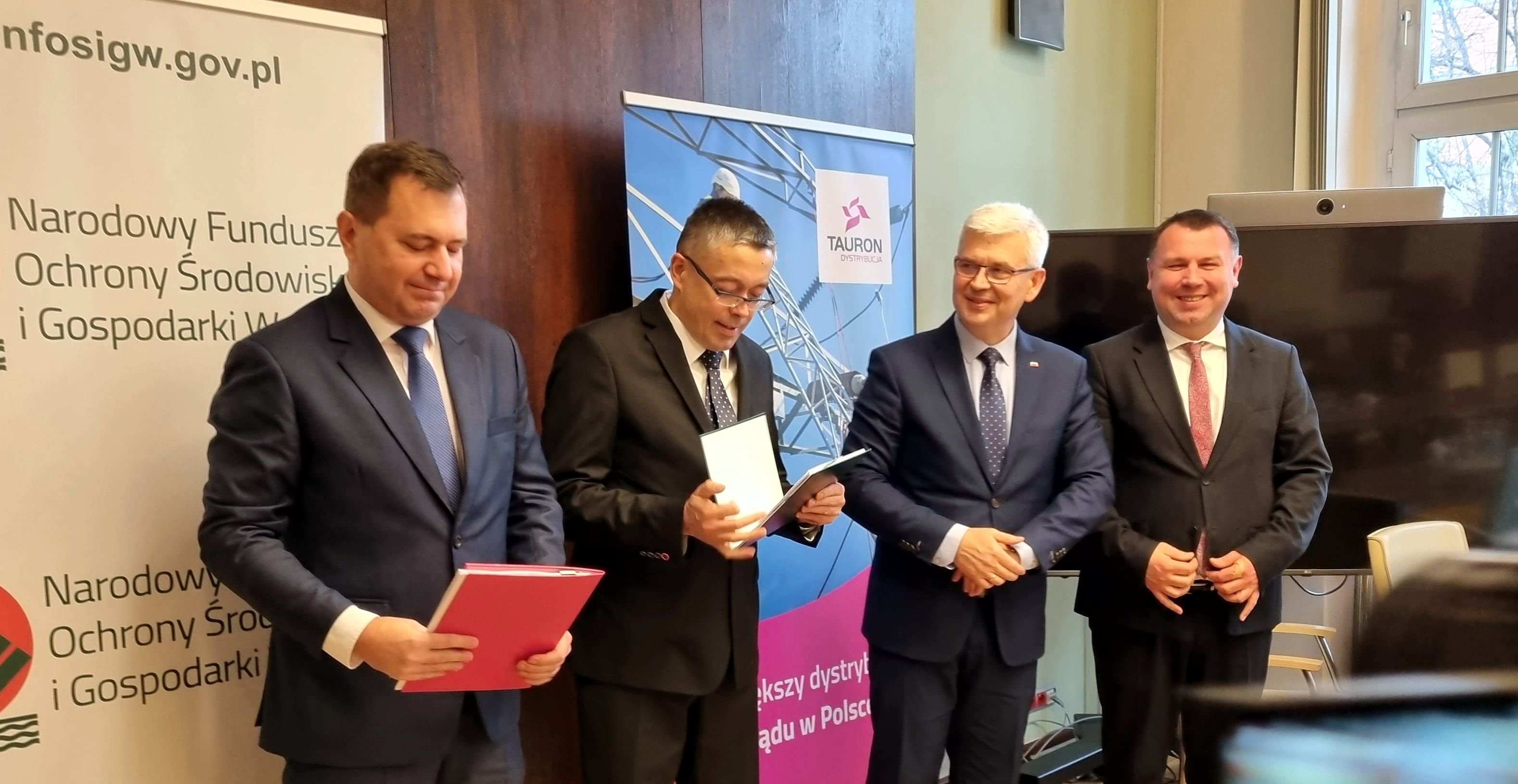 czterech elegancko ubranych i uśmiechniętych panów stoi na tle banerów Narodowego Funduszu Ochrony Środowiska i Gospodarki Wodnej i firmy TAURON Dystrybucja
