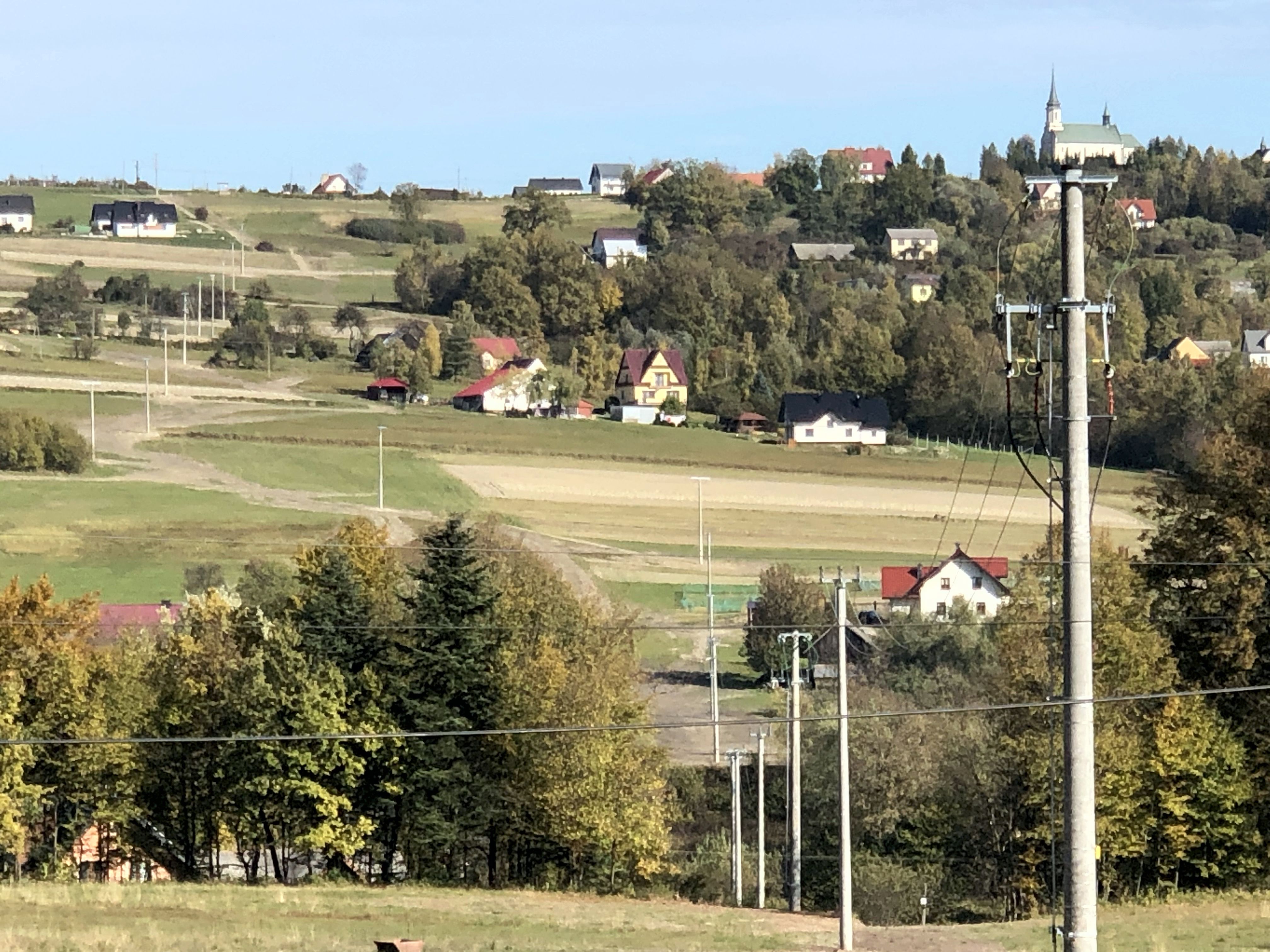 linia energetyczna z wieloma słupami biegnie przez zielony, pagórkowaty krajobraz małopolskiej wsi