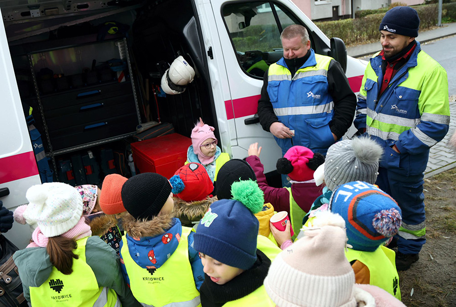 dwóch pracowników pogotowia energetycznego stoi przy samochodzie pogotowia energetycznego i opowiada gromadce dwunastu dzieci - przedszkolaków o swojej pracy