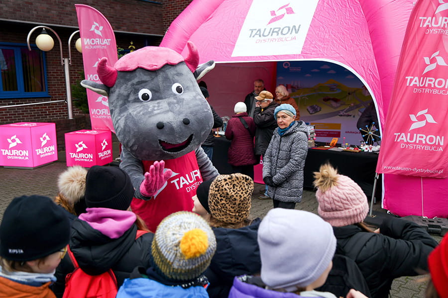 przed magentowym namiotem z logo firmy TAURON Dystrybucja stoi grupka dzieci w wieku wczesnoszkolnym oraz maskotka byczek Tauronek