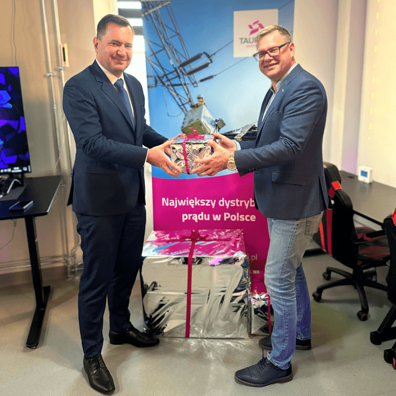 dwóch panów w garniturach trzyma wspólnie w rękach srebrną paczkę z prezentem