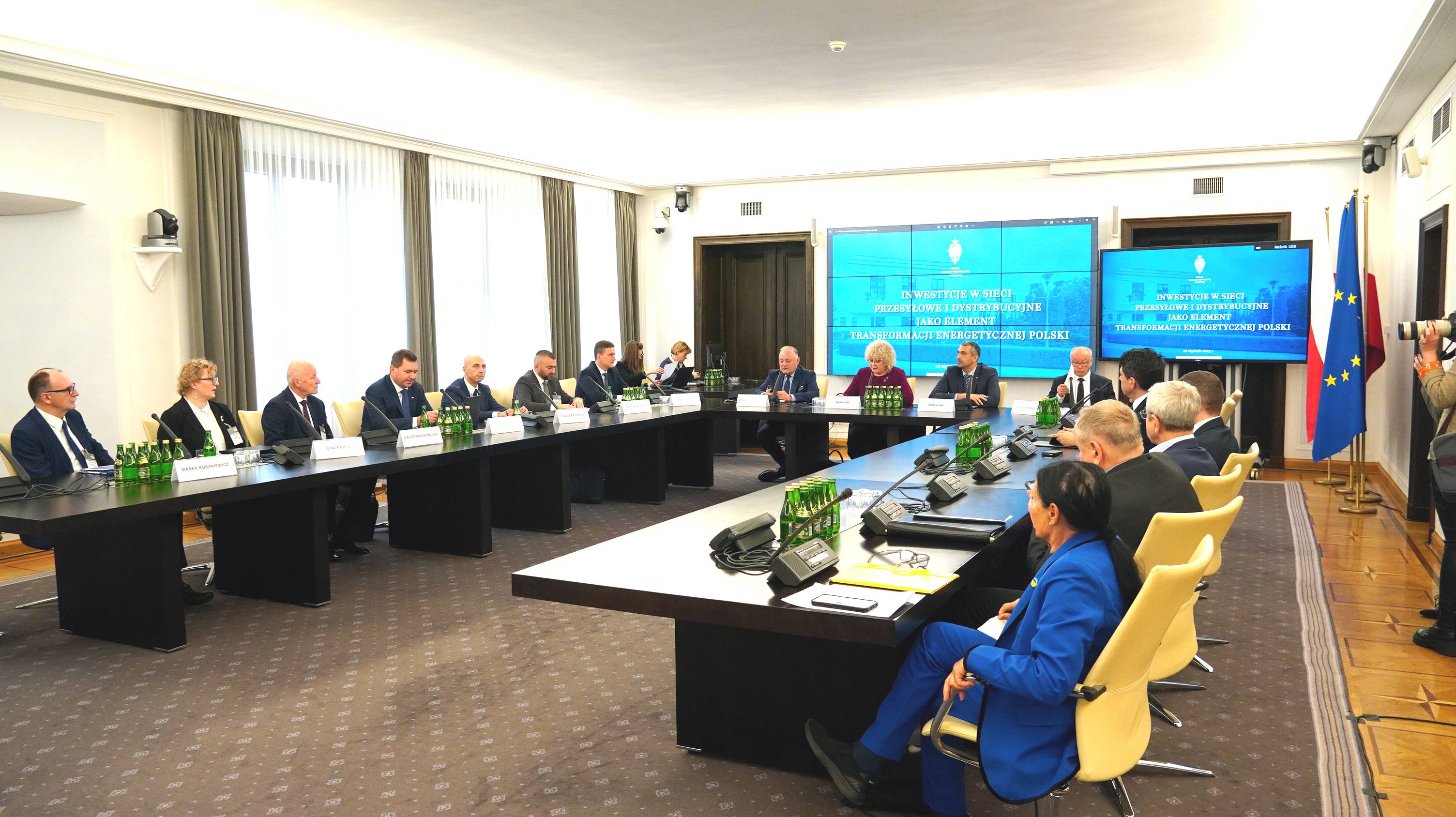 widok na dużą salę konferencyjną na której siedzi osiemnaście osób przy stolikach ustawionych w kształcie podkowy