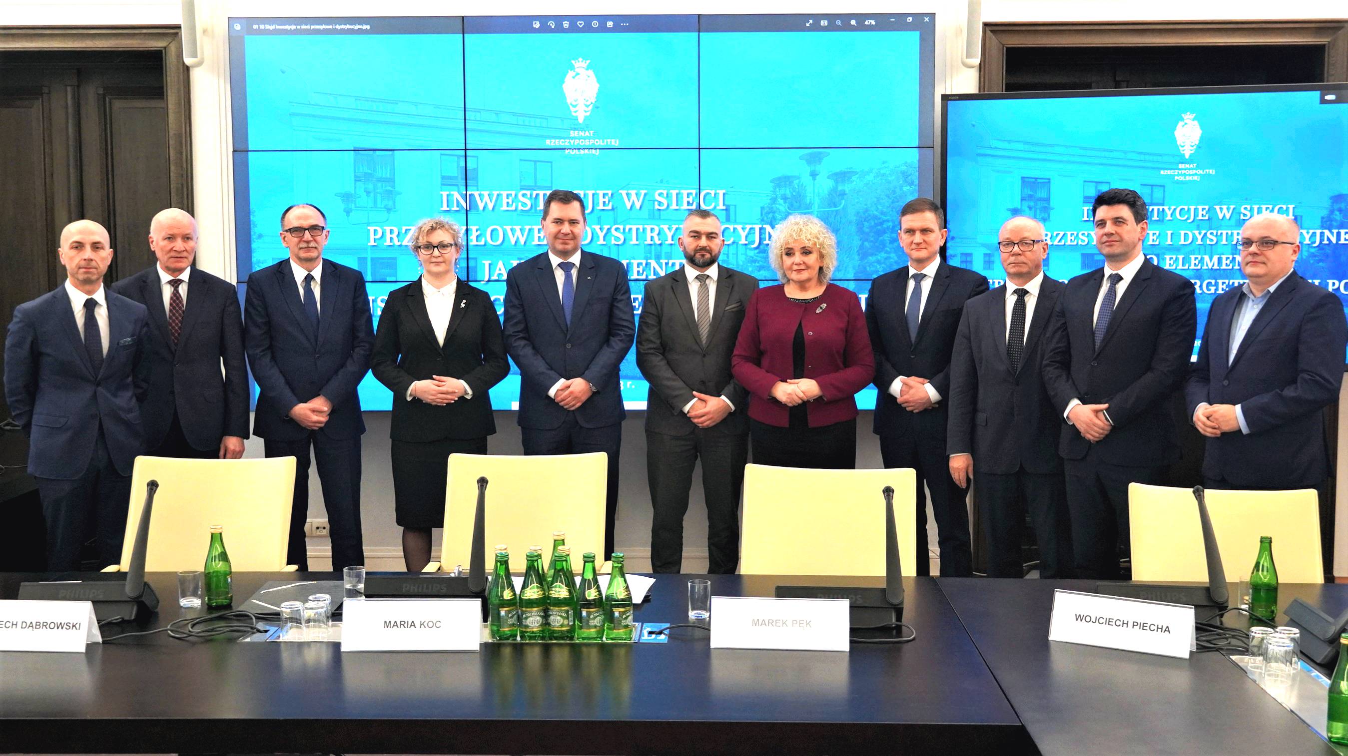  jedenaście elegancko ubranych osób stoi za stolikiem na sali konferencyjnej, za nimi na ekranie wyświetla się tytuł konferencji: Inwestycje w sieci przesyłowe i dystrybucyjne