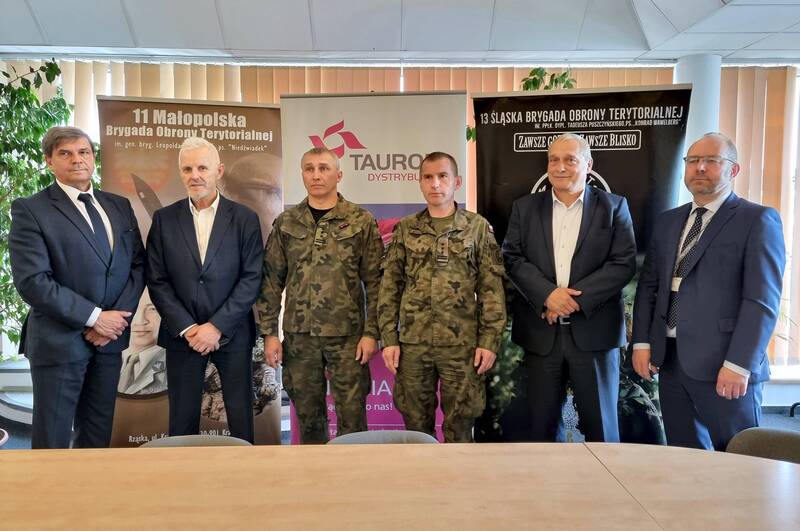 na tle banerów z logo Tauron Dystrybucja i Wojsk Obrony Terytorialnej stoi czterech panów w garniturach i dwóch żołnierzy w mundurach