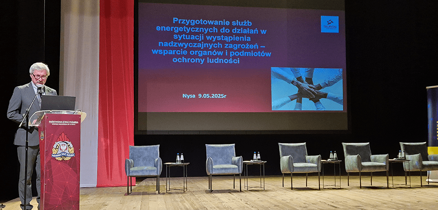 pan ubrany w szary garnitur przemawia przy pulpicie na tle ekranu, na którym wyświetla się tytuł spotkania