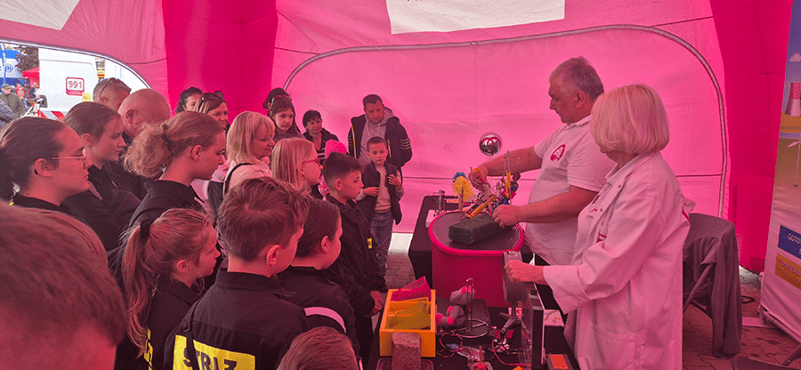 grupa kilkunastu dzieci stoi w magentowym namiocie i przygląda się jak pan i pani prezentują eksperymenty związane z energią elektryczną