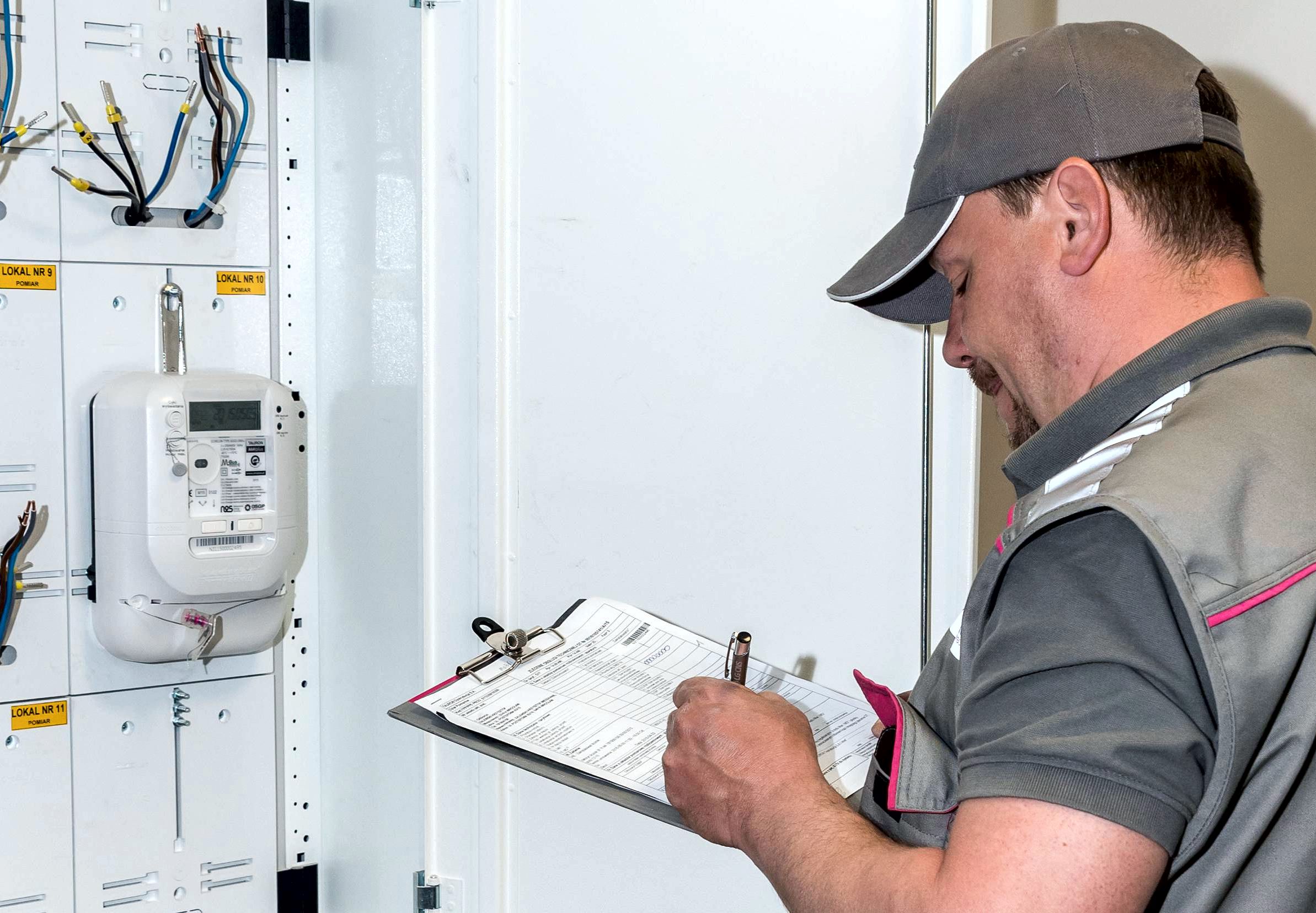 ubrany w szary, służbowy strój przedstawiciel Tauron Dystrybucja zapisuje w notatniku stan licznika, który jest umieszczony na białej szafce licznikowej