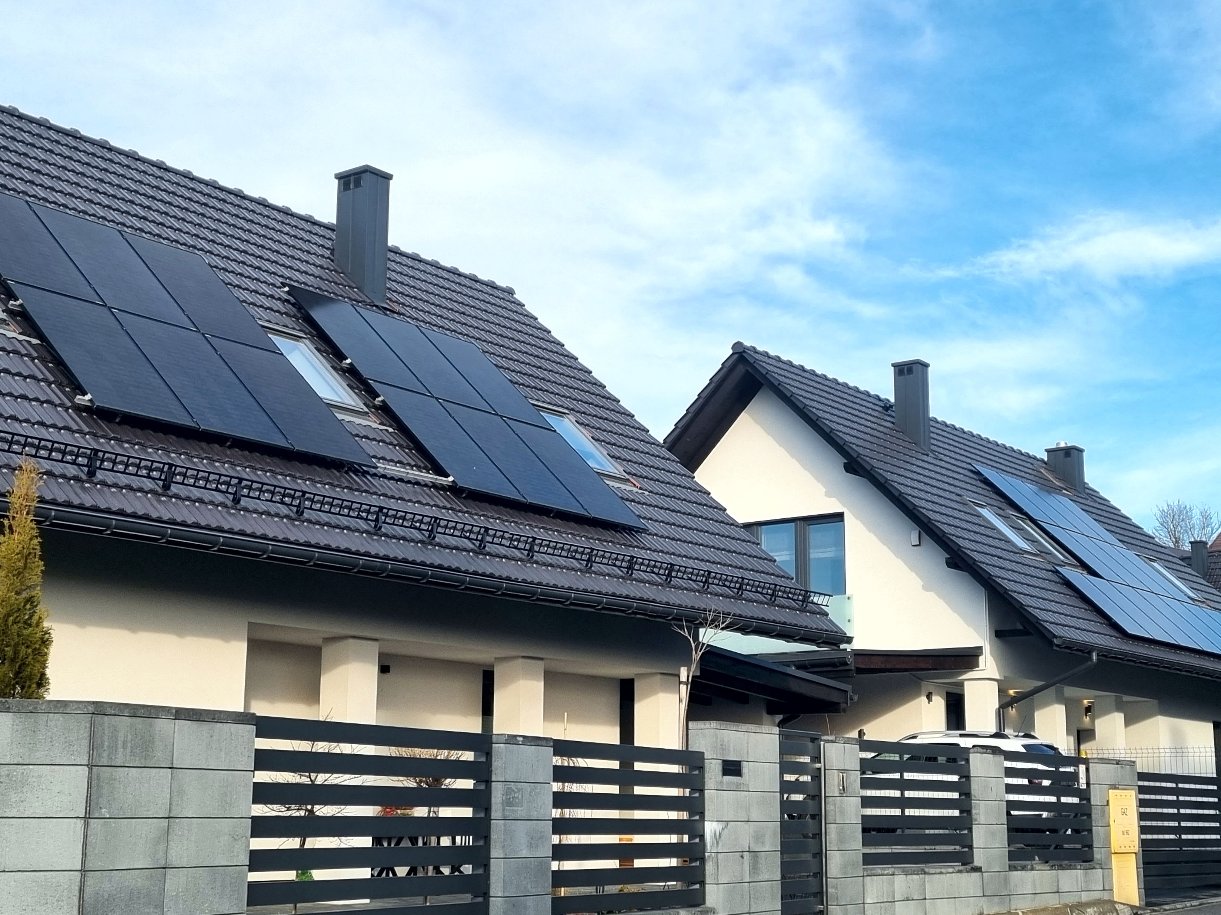 dwa białe domy jednorodzinne, które na szarych dachach mają zamontowane panele fotowoltaiczne