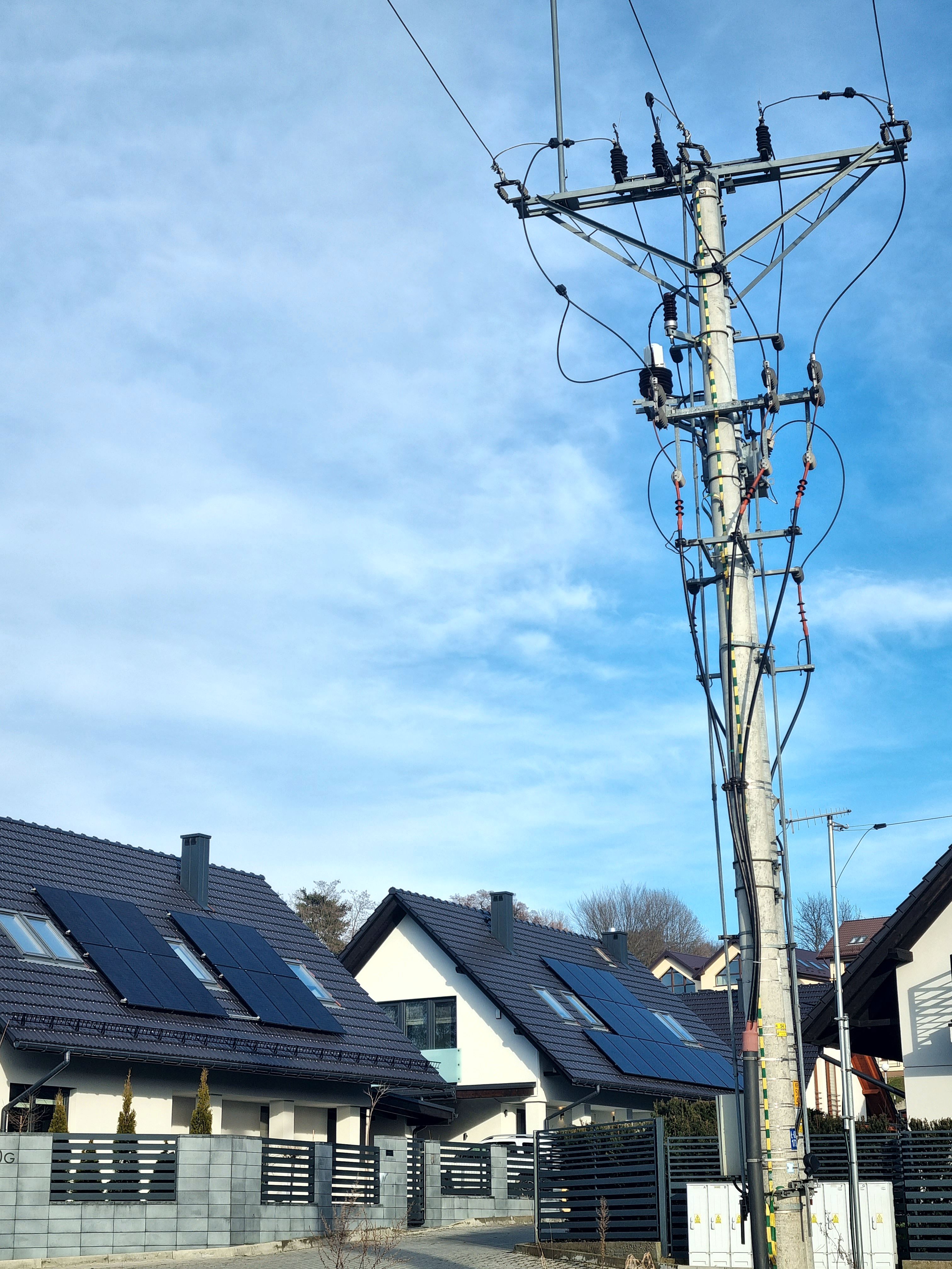 szary słup energetyczny na tle błękitnego nieba oraz osiedla domów jednorodzinnych