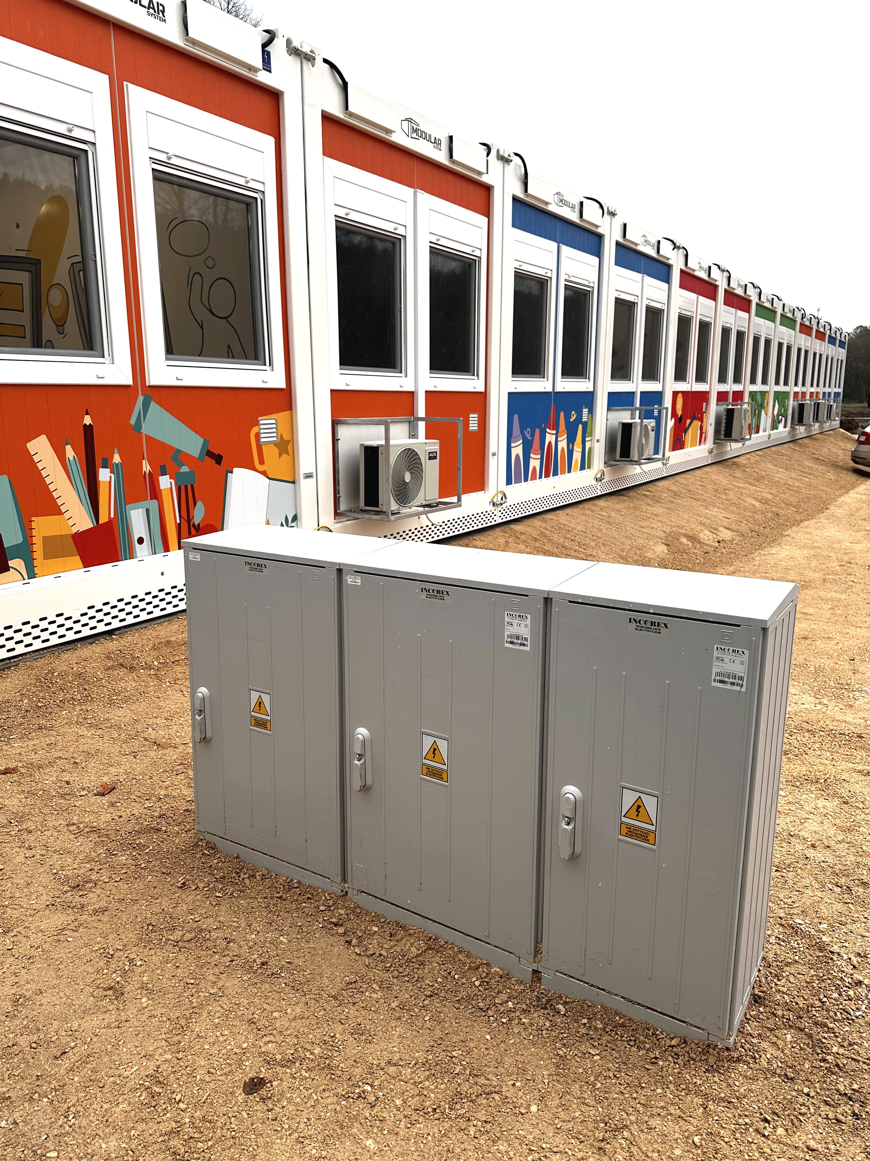 szafki złącz kablowych stojące przed szkołą zbudowaną z kolorowych modułów kontenerowych