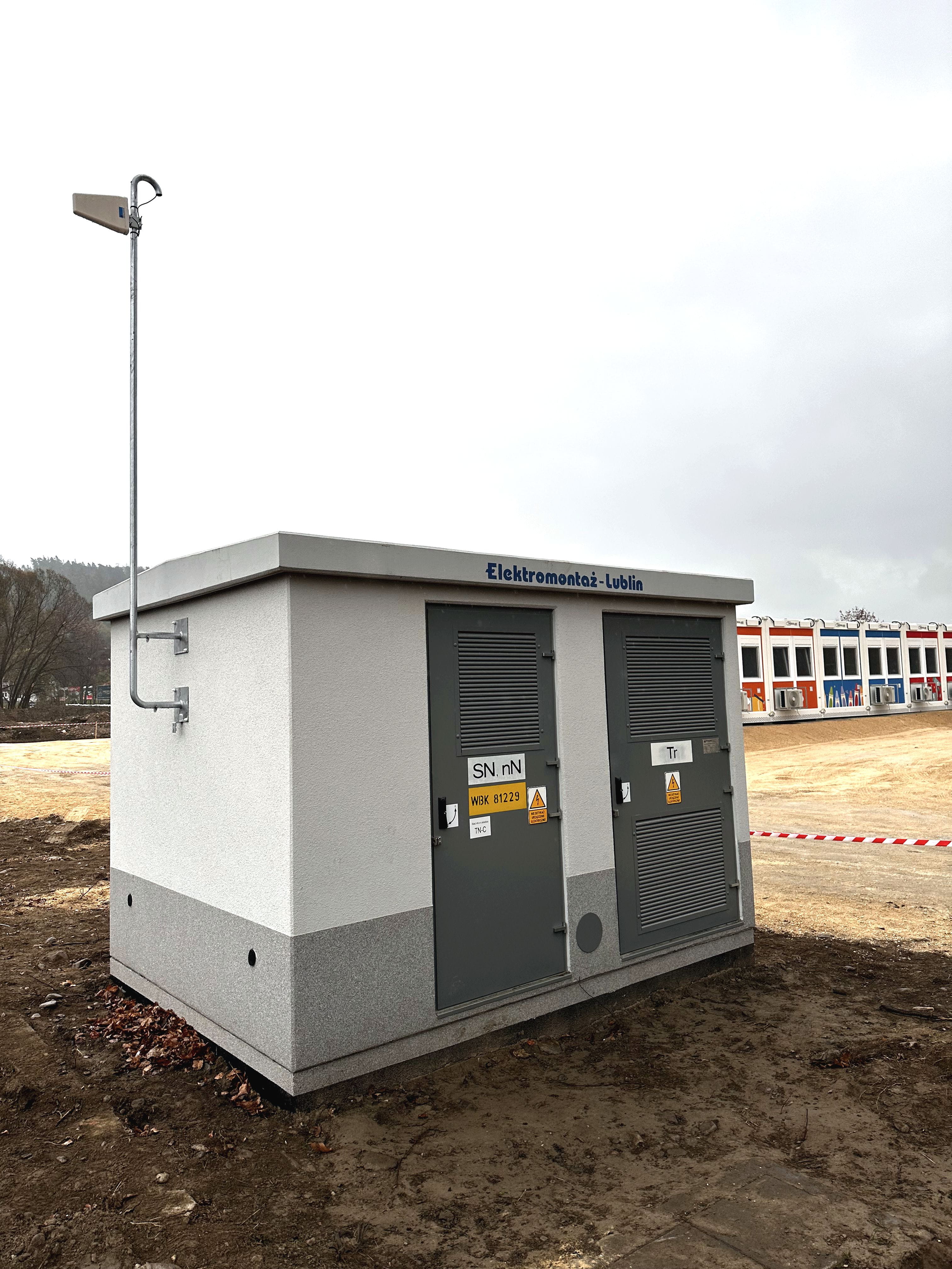 budynek stacji transformatorowej stojący przed szkołą zbudowaną z kolorowych modułów kontenerowych