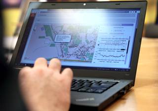 ręka mężczyzny położona jest na klawiaturze laptopa na którego ekranie wyświetla się mapa terenu z zaznaczonymi na niej budynkami